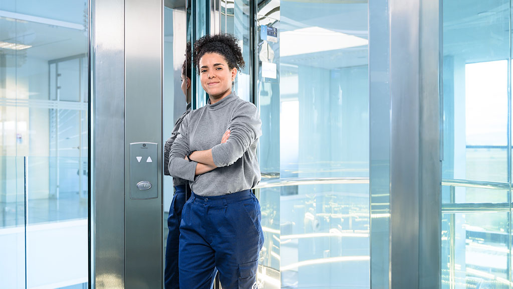 A woman stands in front of a glass lift and looks confidently into the camera.