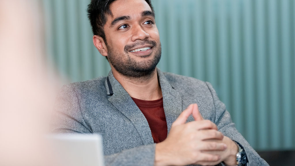 A person in a gray blazer and red shirt sits with hands clasped.