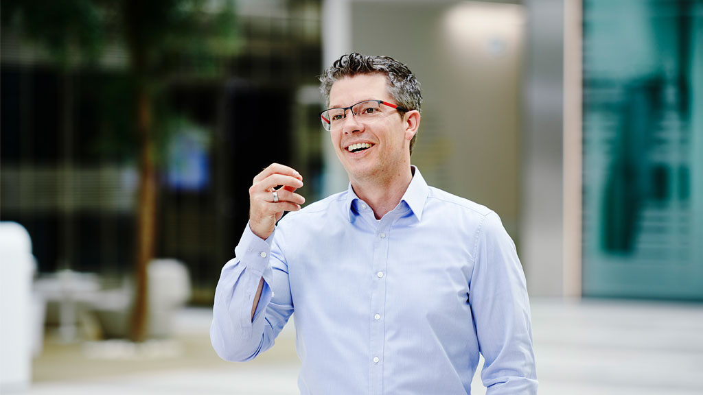 A man stands in the reception area of an office, smiling and gesturing at the camera.