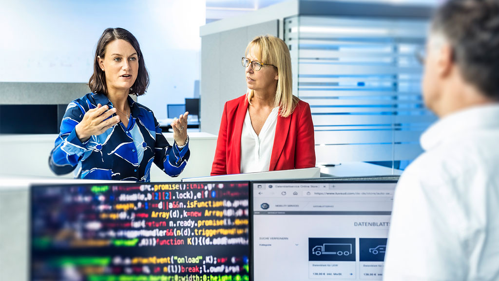 Three people are engaged in a discussion in an office, with two computer monitors in the foreground displaying colorful lines of code.