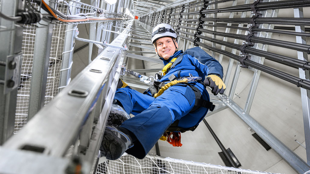 A worker in safety gear, including a helmet and harness, is climbing inside a tall industrial structure, secured with ropes and protective equipment.