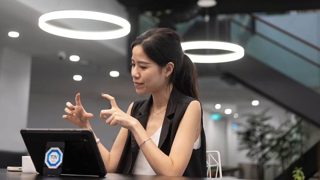 A person sitting at a table in a modern office or meeting space, gesturing with their hands and looking at a tablet device on the table.