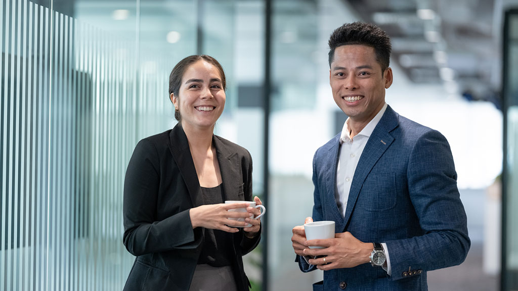 Two individuals stand in an office, holding coffee cups and conversing.