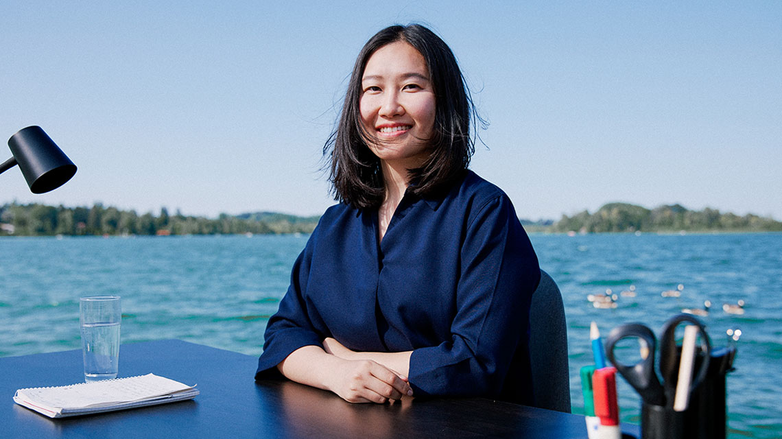 An Asian woman sits at a desk, with a lake in the background.