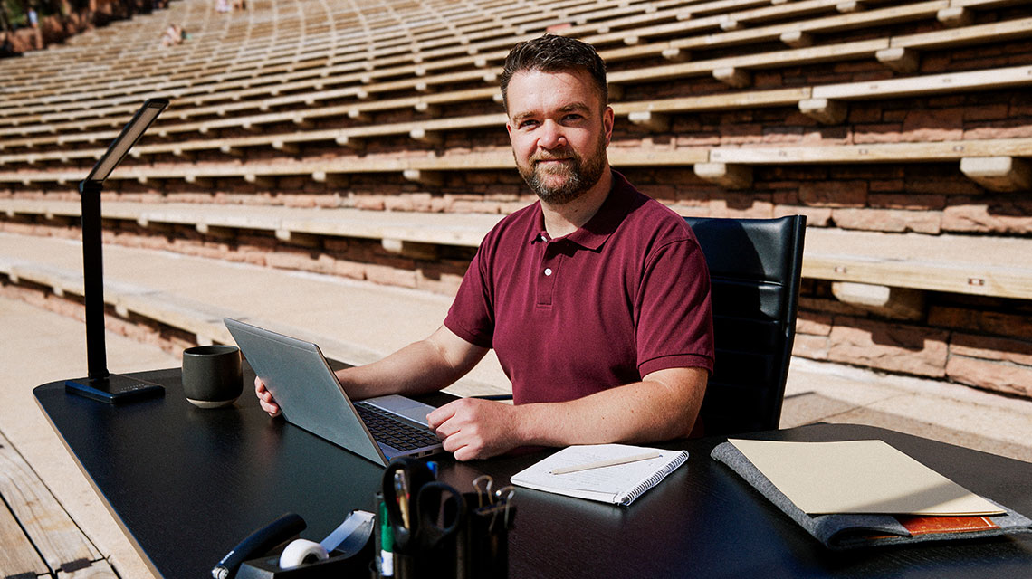 A man sits at a desk, with the stony rows of an amphitheatre in the background.