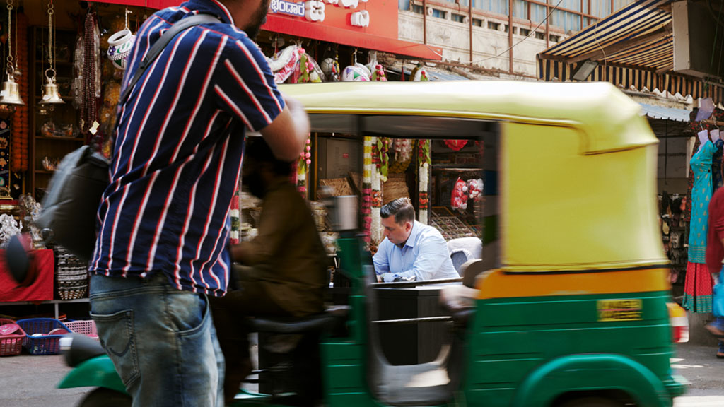 An auto rickshaw drives down a street in India, through the window we see a man sitting at a desk.