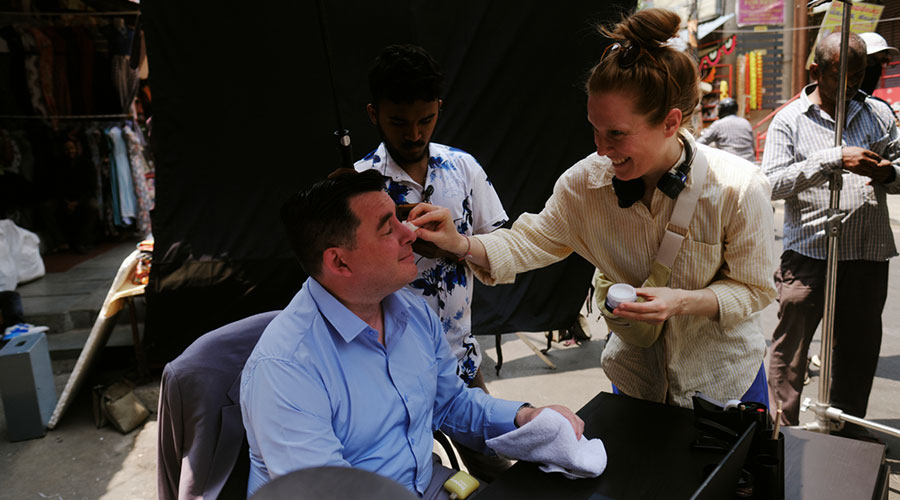 A man is sitting at a desk, a woman is doing his make-up.