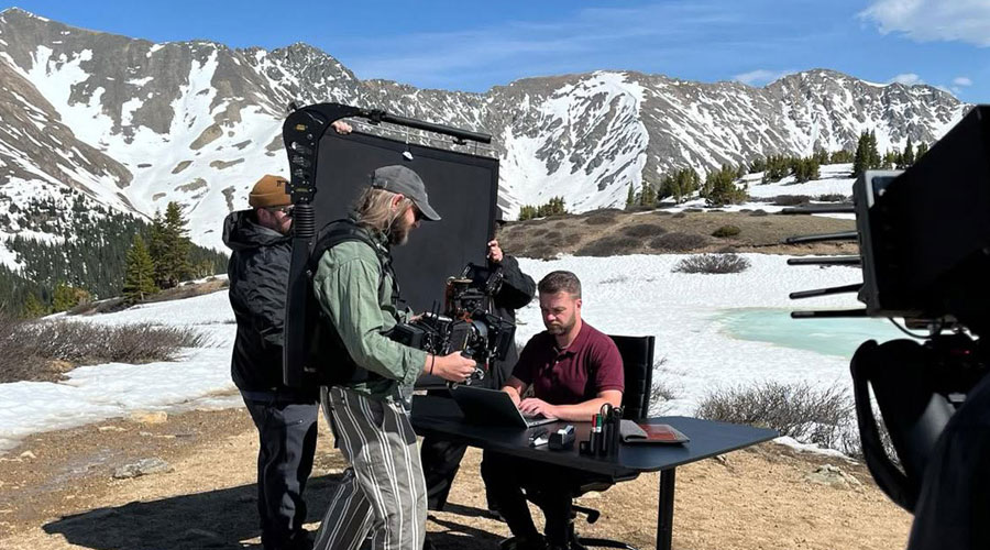 A man is sitting at a desk in a snowy landscape, being filmed by a cameraman.