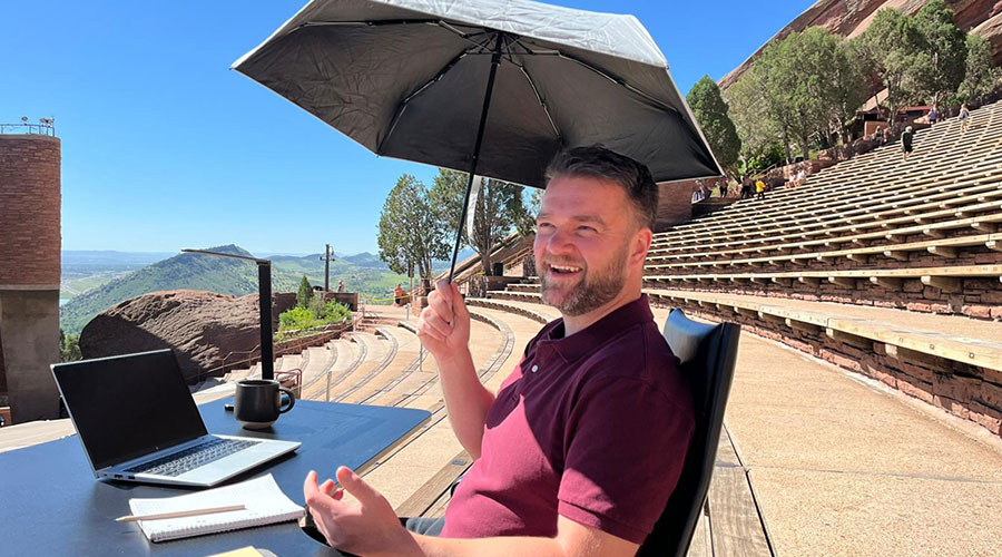 A smiling man sits at a desk in an amphitheatre with an umbrella to protect him from the sun.