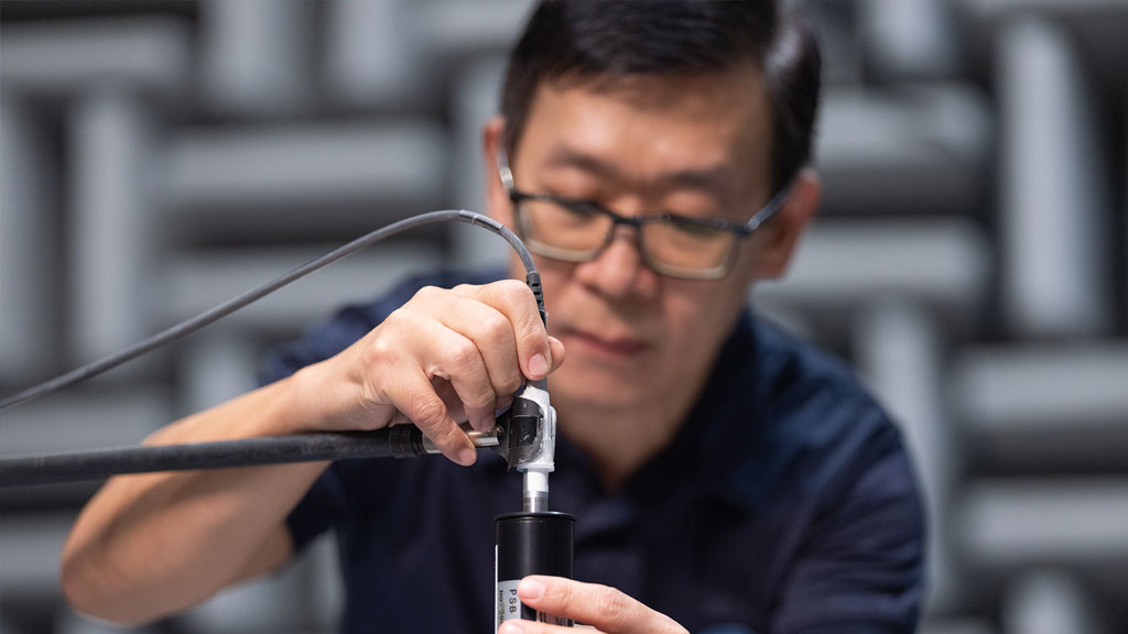 A man wearing glasses and a navy-blue polo shirt is working with a cylindrical device in an acoustic testing chamber.