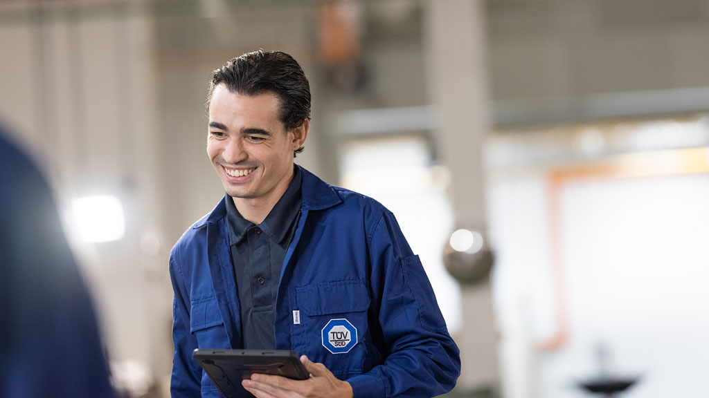 A person wearing a blue jacket with the TÜV SÜD logo on it, holding a tablet. The background appears to be an industrial or laboratory setting with blurred equipment and pipes visible.