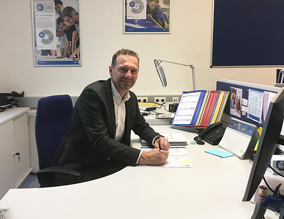 A man is sitting at his desk in his office, smiling into the camera.