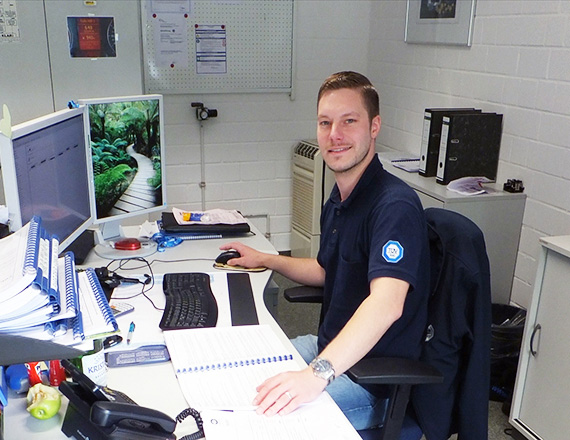 A person wearing a dark blue polo shirt is sitting in front of his computer in his office.