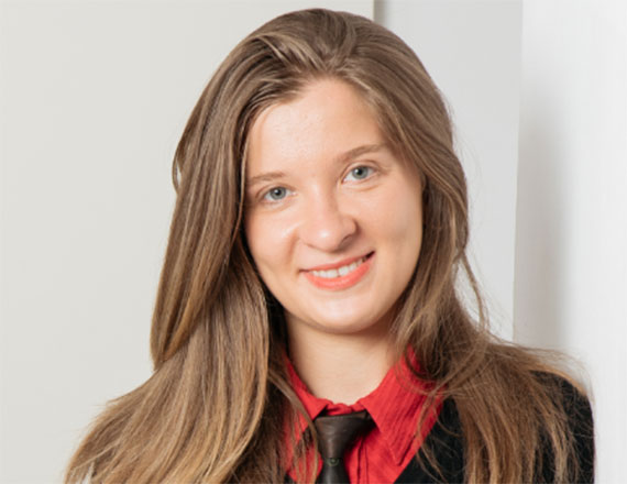 Madalina Burghelea, Head of Data Strategy & Management at TÜV SÜD, is wearing a red shirt, black tie, and black sweater while standing indoors against a white wall, smiling into the camera.
