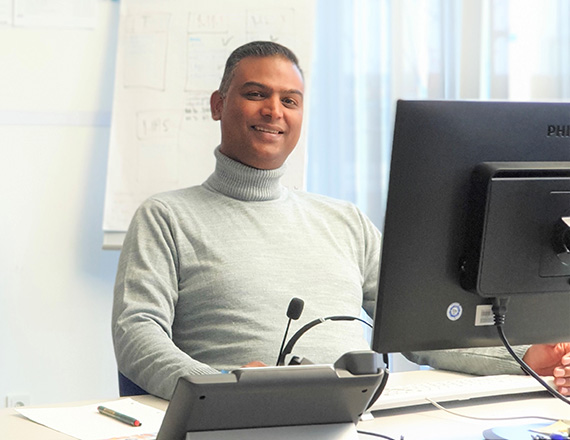 A man in a light grey turtleneck is sitting at his desk in an office.