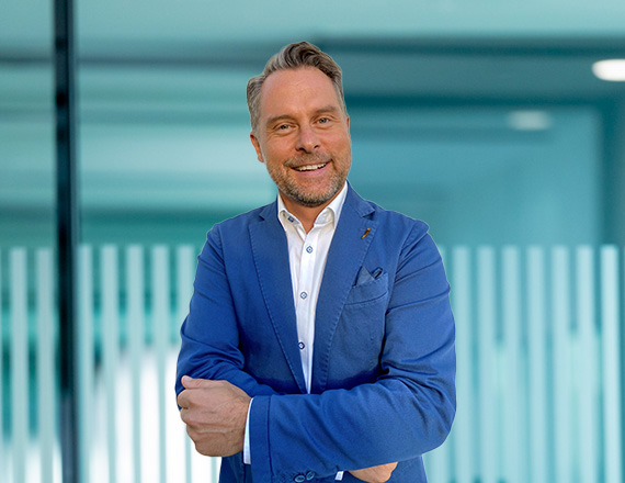 A man in a blue blazer stands in front of a white wall, crosses his arms and smiles at the camera.