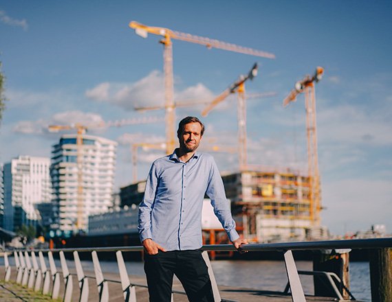 A man in a blue shirt stands on a railing in front of a river, with construction cranes and tall buildings in the background.