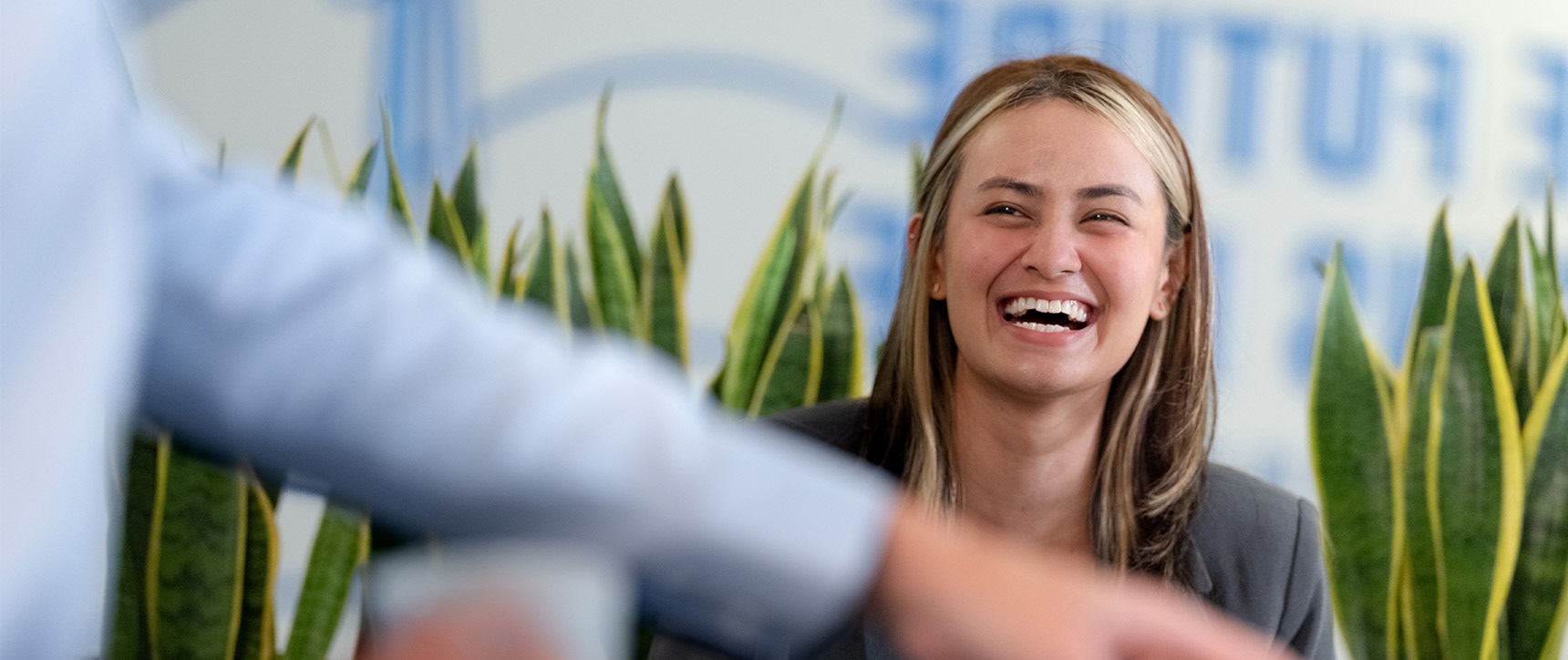 Smiling woman in a business environment. Blurry arm with a finger pointing downwards in the foreground. Plants and a text on the wall saying „The Future“ in the background.
