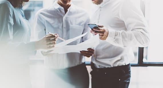 Drei Personen in Businesskleidung stehen zusammen in einem hellen Büro und besprechen Dokumente, während eine Person ein Smartphone hält.