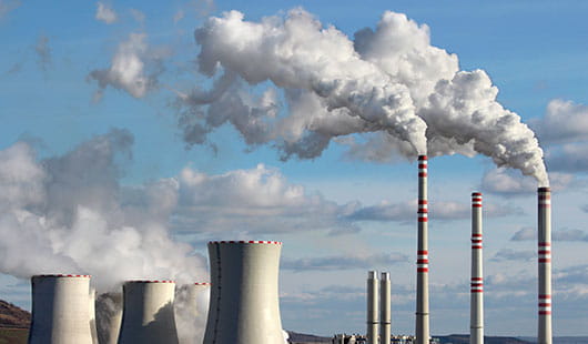 Mehrere Industrie-Schornsteine vor blauem Himmel, aus denen heller Rauch aufsteigt 