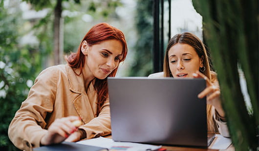 Zwei Frauen sitzen an einem Meeting-Tisch und blicken gemeinsam in ein Laptop