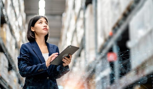 Frau im Business Outfit steht in Hochlager und Blickt auf Bestände. Dabei hält sie ein Tablet in den Händen.