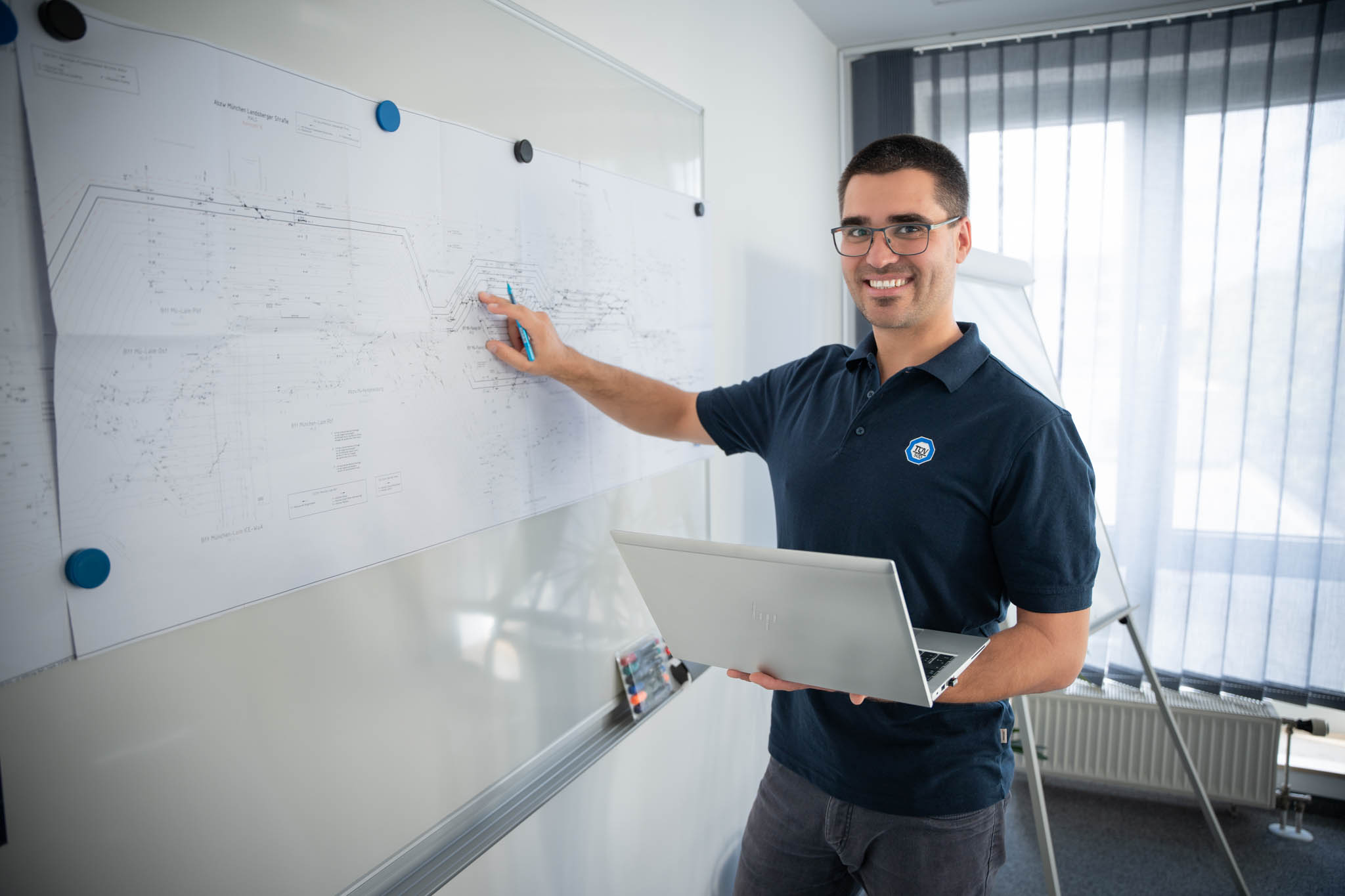 Mann steht mit Laptop in der Hand vor Tafel im Büro und zeigt auf einen Plan.