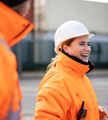 Lachende Frau in Schutzkleidung und Helm auf einer Baustelle.
