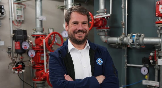 EIn lächelnder TÜV SÜD Angestellter steht vor verschiedenen Rohrleitungen und verschränkt die Arme