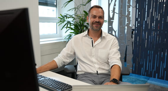 Ein Mann in Arbeitskleidung sitzt vor einem PC und lächelt in die Kamera