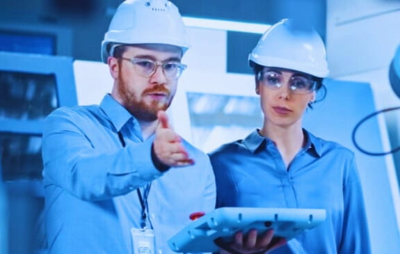 Zwei Ingenieure mit Schutzhelmen überwachen einen Industrieroboter in einer Produktionshalle.