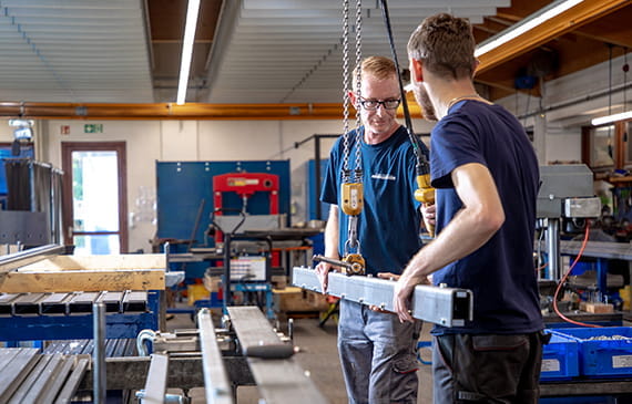 Mitarbeiter in einer Produktionshalle, die gemeinsam eine Hebevorrichtung zur präzisen Montage nutzen.