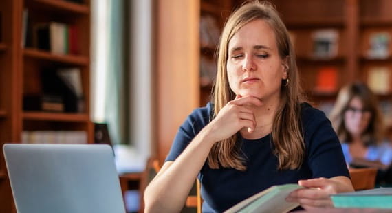 Frau arbeitet konzentriert am Laptop und liest ein Buch in einer Bibliothek, digitale Barrierefreiheit als Thema.