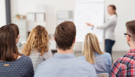 Frau steht vor Gruppe von Personen an Flipchart und pr&auml;sentiert etwas