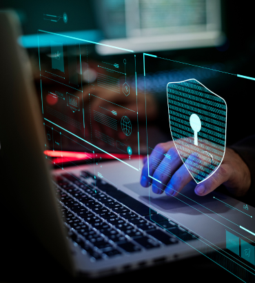 persona che lavora su un laptop, con elementi digitali sovrapposti in realt&agrave; aumentata. In primo piano si nota un simbolo di scudo con una serratura rappresentazione chiara di cybersecurity e protezione dei dati. Lo sfondo presenta elementi grafici digitali, suggerendo monitoraggio, analisi e difesa informatica.