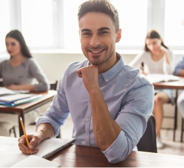 Aula di formazione con alcuni giovani adulti ai banchi. In primo piano, un uomo sorridente con camicia azzurra guarda verso la fotocamera, mentre tiene una matita in mano.